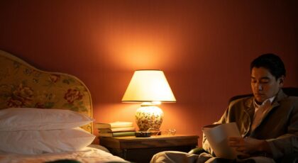 A man sits in a cozy hotel room reading under warm, ambient light.