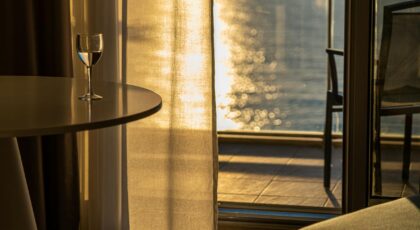 Sunlit room with a wine glass on table, overlooking a serene body of water from a balcony.