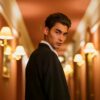 Young man in a black suit standing confidently in a warmly lit hotel corridor.