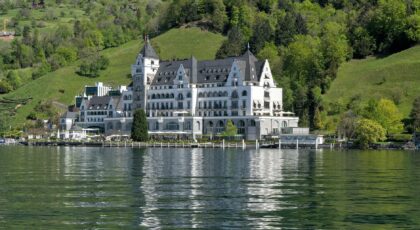 Elegant hotel facade reflecting on a serene Swiss lake with lush green hills in the background.