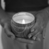 Close-up of a woman holding a lit candle in a patterned container, hands visible.