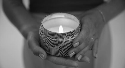 Close-up of a woman holding a lit candle in a patterned container, hands visible.