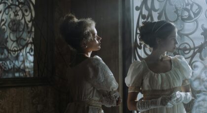 Two women in elegant historical dresses standing by an ornate window indoors.