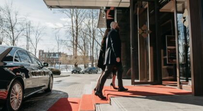 A man and woman elegantly dressed enter a luxury hotel, walking on a red carpet.