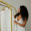 Elegant woman in white dress holding a bathrobe on a gold clothing rack, indoors.
