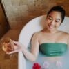 Asian woman enjoying a relaxing bath with wine and flowers, embodying spa-like wellness and tranquility.