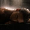 Woman in a spa setting under waterfall, relaxing with eyes closed on a marble surface.