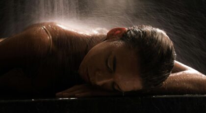 Woman in a spa setting under waterfall, relaxing with eyes closed on a marble surface.